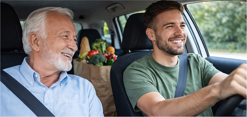 golden era care provider smiles with older male client in passenger seat as they drive home from the grocery store. Healthy groceries are in the backseat.